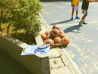 A collection of basketballs is tightly secured in a net, resting on a blue outdoor basketball court. Nearby, two individuals in athletic clothing are partially visible, suggesting a casual or practice environment. A concrete ledge has a clipboard with a basketball strategy diagram, a stopwatch, and a highlighter placed on it. Greenery borders one side of the court, indicating an outdoor setting.