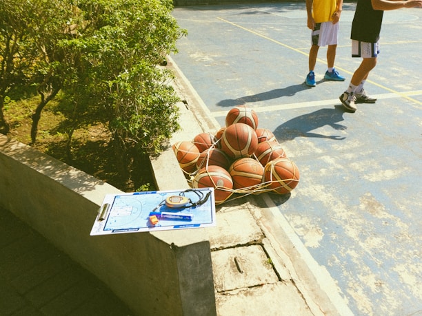 A collection of basketballs is tightly secured in a net, resting on a blue outdoor basketball court. Nearby, two individuals in athletic clothing are partially visible, suggesting a casual or practice environment. A concrete ledge has a clipboard with a basketball strategy diagram, a stopwatch, and a highlighter placed on it. Greenery borders one side of the court, indicating an outdoor setting.