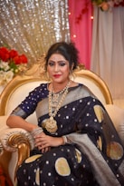 A woman is seated on an ornate gold-trimmed chair. She is dressed in a dark blue saree with gold and silver patterns, complemented by elaborate gold jewelry including a heavy necklace and bracelets. Her hair is styled elegantly, and she wears makeup that highlights her eyes and lips. The background features decorative drapes with lights and floral arrangements.