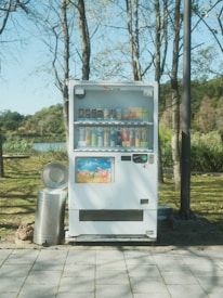 A vending machine is situated outdoors in a park-like setting, surrounded by grass, trees, and a metal trash can. Stacks of colorful drink cans and bottles are visible through the vending machine's glass front.