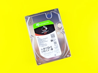 A Seagate IronWolf 8TB internal hard drive is placed against a bright yellow background. The hard drive features a label with the brand name, model, storage capacity, and various specifications and certifications.