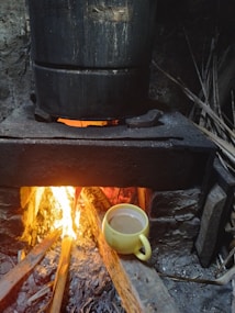 A rustic wood-fired stove with a large metal pot heating over the flames. The fire emits a warm glow, casting light on the surrounding stone and wooden materials. A yellow mug filled with a beverage is placed near the fire, suggesting a cozy, traditional cooking setting.