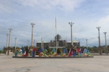 A wide plaza with large, colorful letters spelling 'PUTRAJAYA'. Behind the letters, there are several tall flagpoles and a government building with a dome and flags.