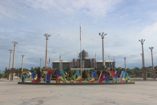 A wide plaza with large, colorful letters spelling 'PUTRAJAYA'. Behind the letters, there are several tall flagpoles and a government building with a dome and flags.