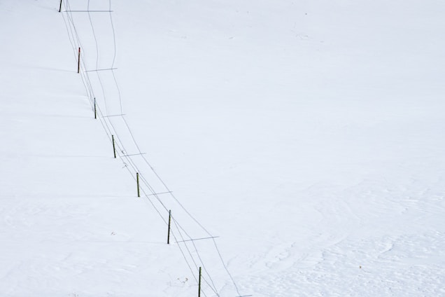 A long fence stretches diagonally across a vast, snow-covered landscape. The fence consists of wooden posts connected by wire.