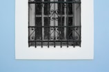 A window with a decorative wrought iron grill set against a smooth blue wall. The grill features intricate scrollwork and patterns, providing a contrast to the simplicity of the wall.