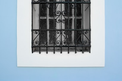 A window with a decorative wrought iron grill set against a smooth blue wall. The grill features intricate scrollwork and patterns, providing a contrast to the simplicity of the wall.