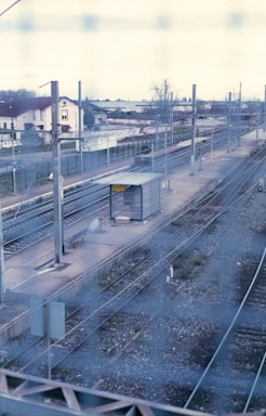A railway platform with several tracks situated in an outdoor setting. There is a small waiting shelter on the platform, surrounded by overhead wires and metal poles. The scene includes residential buildings in the background and is viewed through a metal fence, creating a mesh pattern over the image.