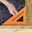 A hand is holding an orange carpenter's square against two pieces of wood, with gray gravel in the background.