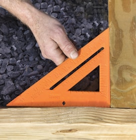 A hand is holding an orange carpenter's square against two pieces of wood, with gray gravel in the background.