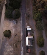 Aerial view of a railway track running vertically. On the right, there's a building complex with mix of metal and wooden roofs, surrounded by trees. Open land with sparse vegetation and dirt roads on the left side.
