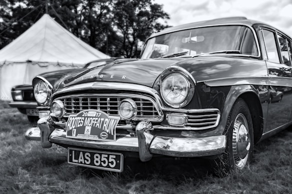A classic vintage car with a chrome grille, round headlights, and a 'Rootes Moffat Run' sign displayed below the front bumper. The car is parked on grass, with a large white tent in the background, partially obscured by trees. The image is rendered in black and white, highlighting the car's shiny exterior and detailed design.