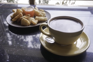 A large cup of coffee is placed on a saucer with a spoon beside it. Next to the coffee, there is a plate of snacks, possibly fried or baked, accompanied by a small glass container of sauce or jam. The background has a reflective surface with a blurred view of trees and sky outside.