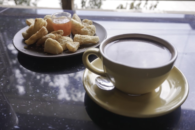 A large cup of coffee is placed on a saucer with a spoon beside it. Next to the coffee, there is a plate of snacks, possibly fried or baked, accompanied by a small glass container of sauce or jam. The background has a reflective surface with a blurred view of trees and sky outside.
