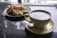 A large cup of coffee is placed on a saucer with a spoon beside it. Next to the coffee, there is a plate of snacks, possibly fried or baked, accompanied by a small glass container of sauce or jam. The background has a reflective surface with a blurred view of trees and sky outside.