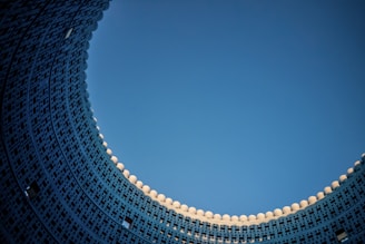 A dynamic architectural structure curves upwards, creating a semi-circular pattern against a clear blue sky. The building features intricate geometric designs and small windows, with a series of white spherical elements crowning the top.