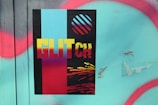A poster features bold text reading 'GLITCH' against a black background divided by vertical stripes in cyan and red. Above the text, a circle with diagonal stripes in cyan and red is depicted. The lower section has a glitch effect with yellow, red, and black colors blending. This is placed on a textured surface with turquoise paint and irregular red spray-painted lines.