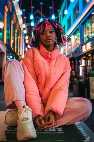 A person sits cross-legged on a platform in a vibrant urban setting. They are wearing a pink puffer jacket, pink pants, and sneakers, with their hair styled in colorful curls. The background is illuminated by various neon lights and fixtures that create a lively atmosphere.