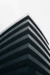 A modern, multi-story building with a minimalist design featuring sharp lines and extensive use of glass and concrete materials. The perspective emphasizes the corner of the structure, creating a sleek and contemporary architectural look against a clear, pale sky.