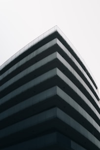 A modern, multi-story building with a minimalist design featuring sharp lines and extensive use of glass and concrete materials. The perspective emphasizes the corner of the structure, creating a sleek and contemporary architectural look against a clear, pale sky.