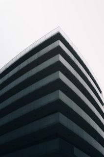 A modern, multi-story building with a minimalist design featuring sharp lines and extensive use of glass and concrete materials. The perspective emphasizes the corner of the structure, creating a sleek and contemporary architectural look against a clear, pale sky.