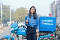 A young woman wearing a blue shirt and black pants is smiling and giving a thumbs-up gesture while standing next to a blue delivery motorcycle with a Domino's branded box attached to the back. The setting appears to be an urban street with trees and buildings visible in the background.