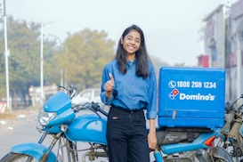 A young woman wearing a blue shirt and black pants is smiling and giving a thumbs-up gesture while standing next to a blue delivery motorcycle with a Domino's branded box attached to the back. The setting appears to be an urban street with trees and buildings visible in the background.