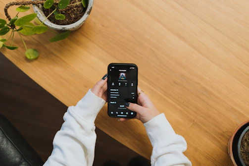 A person wearing a white long-sleeve shirt is holding a smartphone, displaying a music application. The scene is set on a wooden table with a potted plant visible in the corner.