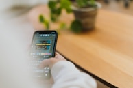 A person is using a smartphone, likely engaged with a social media app or similar, as suggested by a text reading 'DOWNLOAD HERE' displayed on the screen. The background features a wooden surface, possibly a table or desk, with a potted plant and a blurred out setting.