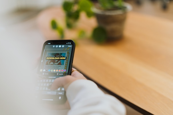 A person is using a smartphone, likely engaged with a social media app or similar, as suggested by a text reading 'DOWNLOAD HERE' displayed on the screen. The background features a wooden surface, possibly a table or desk, with a potted plant and a blurred out setting.