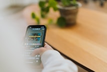 A person is using a smartphone, likely engaged with a social media app or similar, as suggested by a text reading 'DOWNLOAD HERE' displayed on the screen. The background features a wooden surface, possibly a table or desk, with a potted plant and a blurred out setting.
