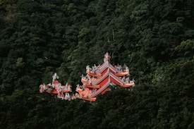 A traditional Asian temple with ornate red and orange rooftops is nestled amidst lush green forest trees.