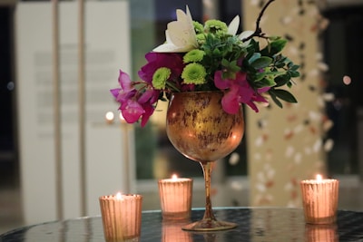 A rustic golden chalice filled with a vibrant arrangement of flowers, including green chrysanthemums and pink bougainvillea, is centered on a reflective table. Surrounding the chalice are three small lit candles in textured golden holders, creating a warm and intimate ambiance.