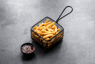 A serving of crispy golden French fries is presented in a black metal basket. Next to it, a small black bowl contains a dark red dipping sauce garnished with green herbs. The setting is on a gray textured surface, creating a modern and minimalistic look.