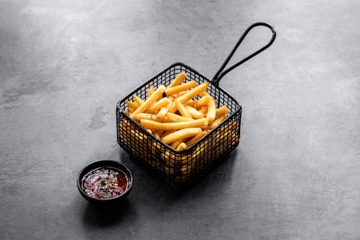 A serving of crispy golden French fries is presented in a black metal basket. Next to it, a small black bowl contains a dark red dipping sauce garnished with green herbs. The setting is on a gray textured surface, creating a modern and minimalistic look.