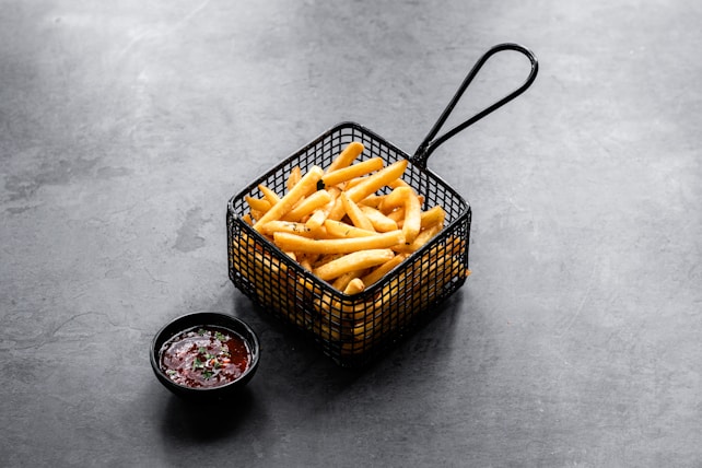 A serving of crispy golden French fries is presented in a black metal basket. Next to it, a small black bowl contains a dark red dipping sauce garnished with green herbs. The setting is on a gray textured surface, creating a modern and minimalistic look.