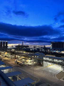 A large industrial complex is visible, with various structures, tanks, and buildings illuminated under a darkening blue sky with scattered clouds. Streetlights cast an artificial glow on the asphalt roads and pathways of the facility.
