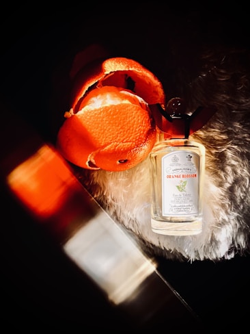 A bottle of Penhaligon's Orange Blossom fragrance lies on a soft, textured surface next to an opened orange peel. The scene is warmly lit with orange and gold hues, casting dramatic shadows.