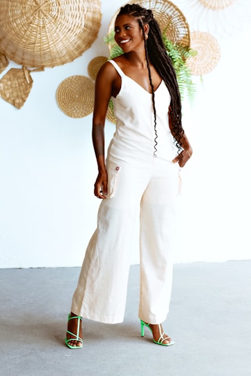 A person with long braided hair and a white jumpsuit is standing confidently. They are wearing bright green strappy high-heeled sandals against a backdrop of woven wall decorations and a few green plants.