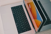 Two laptops are placed side by side on a light-colored wooden surface. One laptop is partially open, and the other is lying flat. The open laptop's screen reflects bright orange, blue, and yellow hues, suggesting a vibrant graphical display. Both laptops have black keyboards.