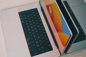 Two laptops are placed side by side on a light-colored wooden surface. One laptop is partially open, and the other is lying flat. The open laptop's screen reflects bright orange, blue, and yellow hues, suggesting a vibrant graphical display. Both laptops have black keyboards.