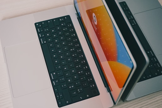 Two laptops are placed side by side on a light-colored wooden surface. One laptop is partially open, and the other is lying flat. The open laptop's screen reflects bright orange, blue, and yellow hues, suggesting a vibrant graphical display. Both laptops have black keyboards.