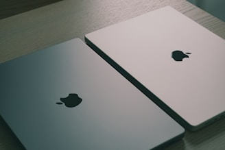 Two closed laptops with the Apple logo are placed side by side on a light wooden surface. One laptop has a gray finish, and the other has a light silver finish, both reflecting the ambient light softly.