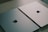 Two closed laptops with the Apple logo are placed side by side on a light wooden surface. One laptop has a gray finish, and the other has a light silver finish, both reflecting the ambient light softly.