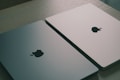 Two closed laptops with the Apple logo are placed side by side on a light wooden surface. One laptop has a gray finish, and the other has a light silver finish, both reflecting the ambient light softly.