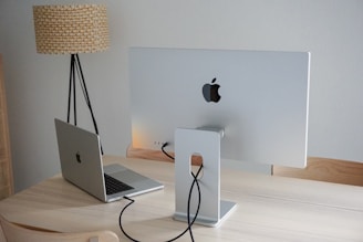 A modern workspace featuring a silver desktop monitor and laptop, both with a recognizable logo, are placed on a light wooden desk. The monitor's back is visible, showing its stand and connected cables. A tall floor lamp with a textured shade stands beside the desk against a plain white wall.