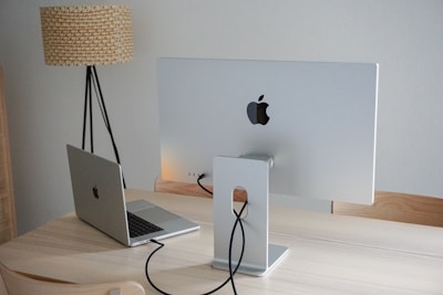 A modern workspace featuring a silver desktop monitor and laptop, both with a recognizable logo, are placed on a light wooden desk. The monitor's back is visible, showing its stand and connected cables. A tall floor lamp with a textured shade stands beside the desk against a plain white wall.