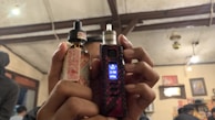 A person is holding a vaping device in their right hand and a bottle of e-liquid in their left hand. The background features an indoor setting with visible wooden ceiling beams, some lit hanging lights, wall decorations including pictures or posters, and other people present.