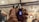 A person is holding a vaping device in their right hand and a bottle of e-liquid in their left hand. The background features an indoor setting with visible wooden ceiling beams, some lit hanging lights, wall decorations including pictures or posters, and other people present.