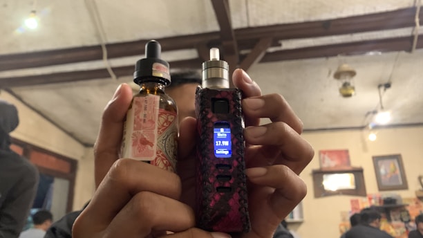 A person is holding a vaping device in their right hand and a bottle of e-liquid in their left hand. The background features an indoor setting with visible wooden ceiling beams, some lit hanging lights, wall decorations including pictures or posters, and other people present.
